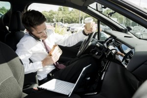 A man in business attire multitasking in a car, holding a coffee cup and looking at his phone with a laptop open beside him.