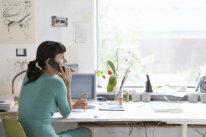 A woman sitting at a desk in a bright office, talking on a mobile phone while looking at a computer screen.