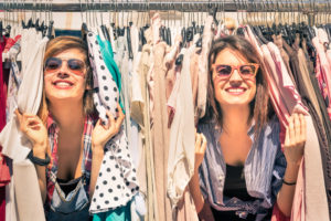 Two smiling women wearing sunglasses peek through rows of clothing on a rack.