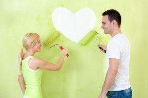 A smiling couple painting a white heart on a green wall with rollers.