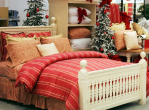 A festively decorated bedroom display with a christmas theme, featuring a bed with red and gold bedding and pillows, flanked by christmas trees.