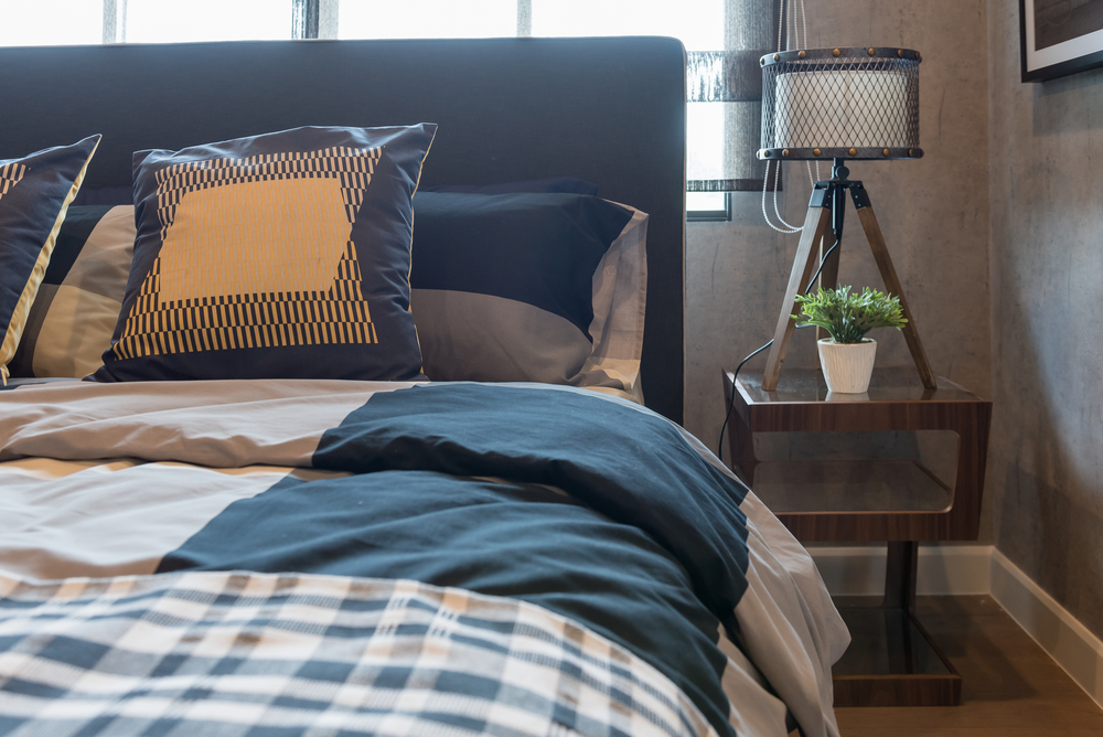 A modern bedroom featuring a neatly made bed with plaid bedding and decorative pillows, adjacent to a wooden bedside table with a lamp and a small plant.