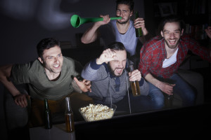Four men cheering and watching a sports game on tv at night with drinks and snacks.