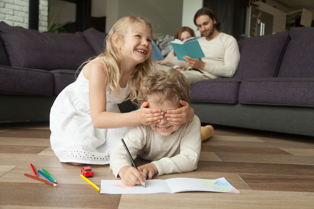 Young girl playfully covering a boy's eyes while he draws, with two adults reading in the background.