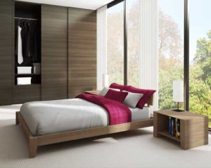 Modern bedroom with a wooden bed frame, white and red bedding, and floor-to-ceiling windows overlooking a wooded area.