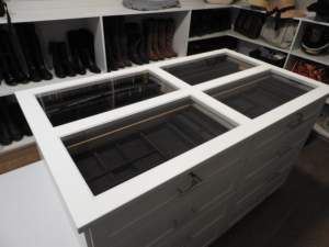 A well-organized closet featuring a white storage island with glass-top drawers and a variety of shoes and boots on shelves.