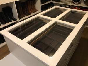 A white storage unit with transparent top compartments, situated in a room with a collection of boots on shelves in the background.