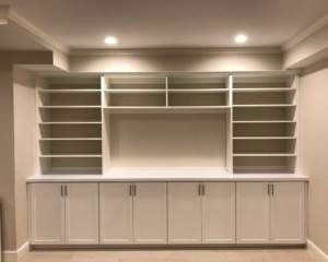 A newly constructed, empty white built-in shelving unit and cabinets against a wall with recessed lighting.