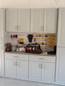 A garage workshop area with white cabinets, a pegboard with hanging tools, and a workbench with various tools and supplies.