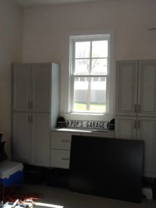 A room corner with white cabinets, a decorative sign reading "pop's garage," a window with a view of outside, and a large black flat screen on a desk.