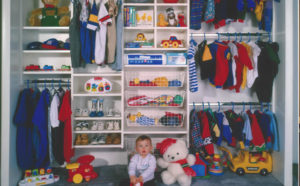 A child sits on the floor of a neatly organized closet filled with toys, clothes, and stuffed animals.