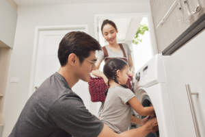 A family doing laundry together with the father loading the washing machine as the mother and daughter watch.