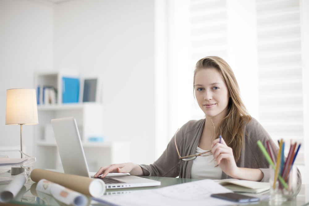 A woman working at a desk with a laptop and papers, looking towards the camera.