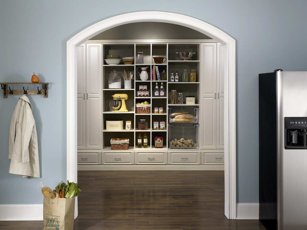 A well-organized kitchen pantry with an arched design, featuring neatly arranged shelves with food items and kitchenware, adjacent to a stainless steel refrigerator.