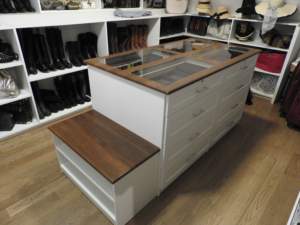 A well-organized closet featuring a central island with drawers and a glass top, surrounded by shelves of shoes and boots.