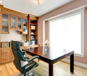 A well-lit home office with a large desk, leather chair, and built-in wooden shelving.
