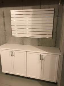 A white storage cabinet with doors sits beneath a horizontal, slatted wall decoration mounted on a bare concrete wall.
