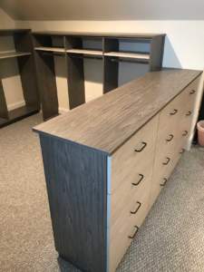 A wooden drawer cabinet in front of an empty bookshelf in a carpeted room.