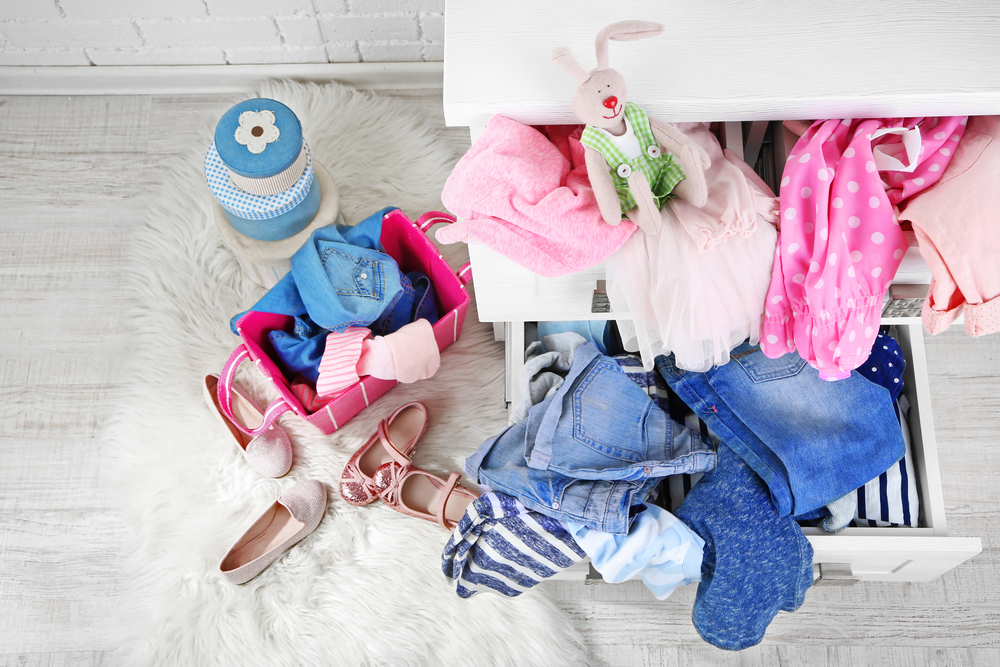 A drawer filled with children's clothing items and a toy rabbit, with additional clothes and shoes scattered on a white fluffy rug.