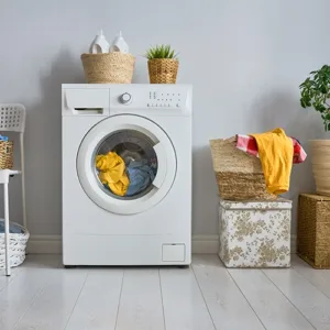 Front-loading washing machine with laundry inside, flanked by wicker baskets and potted plants in a clean, bright custom closet.