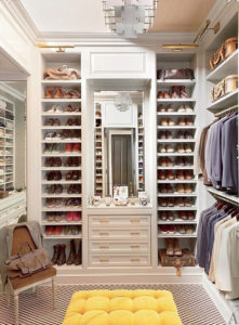 An elegantly designed walk-in closet with neatly organized shoes, clothing, and accessories, featuring a central full-length mirror and a plush yellow ottoman.