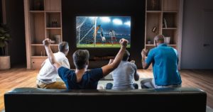 Four people on a couch raising their arms in excitement while watching a soccer match on a large television in a home setting.