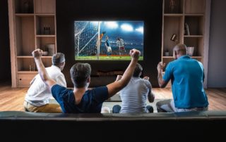 Four people on a couch raising their arms in excitement while watching a soccer match on a large television in a home setting.