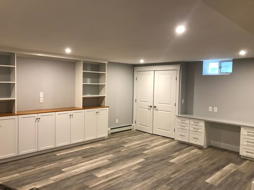 A modern empty basement room with custom entertainment centers, built-in white cabinets, and grey flooring.