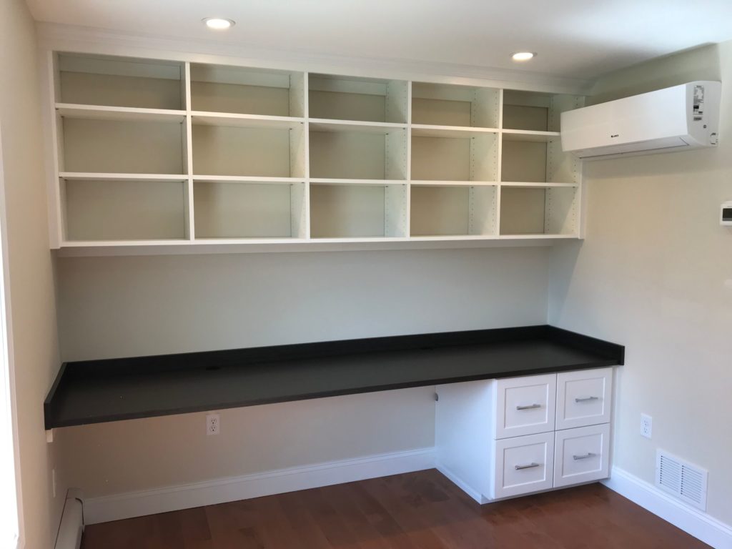 An empty white built-in desk with overhead shelving and white drawers in a clean office room.