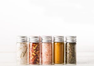 Five spice jars with various seasonings aligned on a light background.