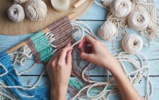 Person weaving a macramé wall hanging with various colors of yarn on a wooden table.