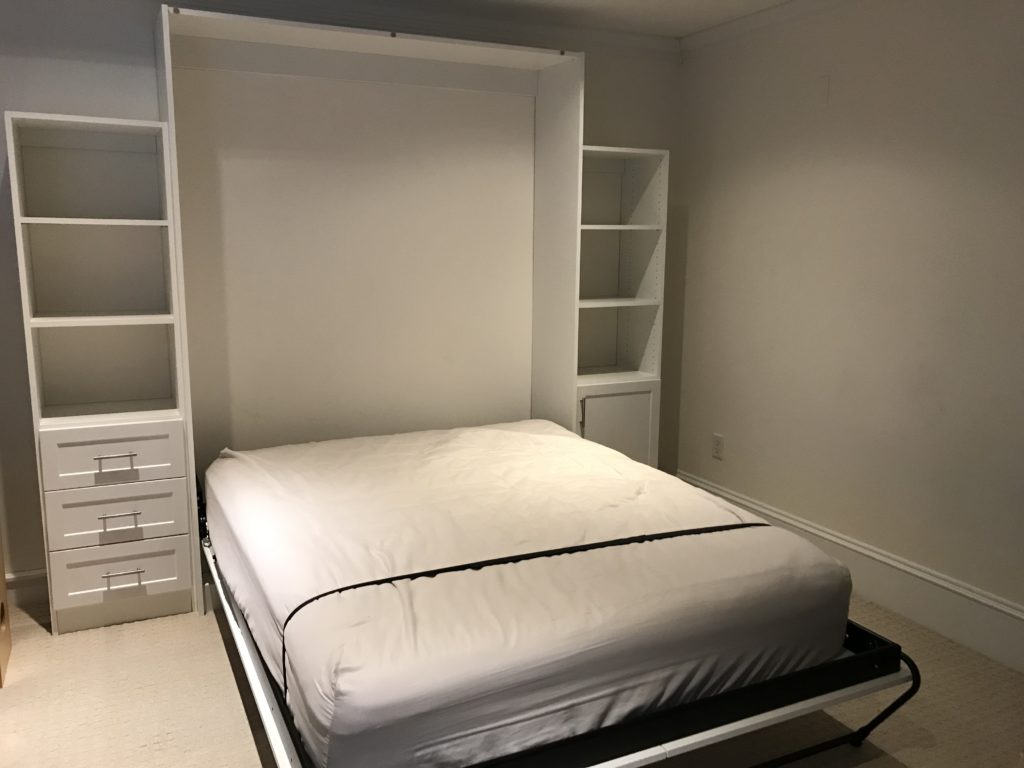 A murphy bed partially folded down in a clean room with built-in shelves and drawers.