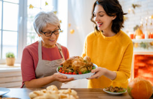 Two women, one older with gray hair wearing a red apron and one younger in a yellow sweater, smiling as they prepare to serve a roasted chicken on a dining table decorated for a festive occasion.