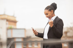 Professional woman in work clothes smiling and looking at her smartphone, displaying a gesture of success or excitement.