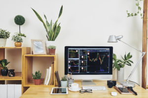 A well-organized home office workstation with a computer displaying stock market charts, surrounded by plants, books, and photography equipment.