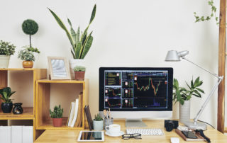 A well-organized home office workstation with a computer displaying stock market charts, surrounded by plants, books, and photography equipment.