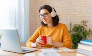 A smiling woman wearing headphones and glasses, holding a red mug while looking at her laptop in a well-lit home office design.