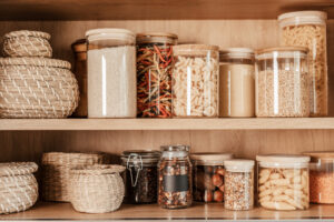 A neatly organized pantry shelf containing various dry food items in clear glass jars and woven baskets.