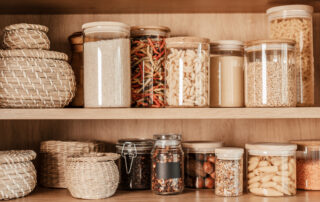 A neatly organized pantry shelf containing various dry food items in clear glass jars and woven baskets.