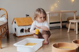 Toddler playing with dry pasta on a wooden floor next to a Murphy Bed indoors.
