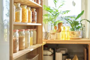 Assorted dry food items stored in clear jars on wooden pantry shelves, with sunlight streaming in.