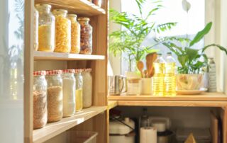 Assorted dry food items stored in clear jars on wooden pantry shelves, with sunlight streaming in.