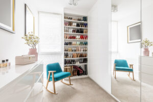 Modern walk-in closet with a neatly organized shoe storage area, a mirrored wardrobe, and a seating area with turquoise chairs.