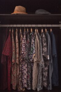An assortment of patterned shirts hanging in a reach-in closet with two hats placed on a shelf above.