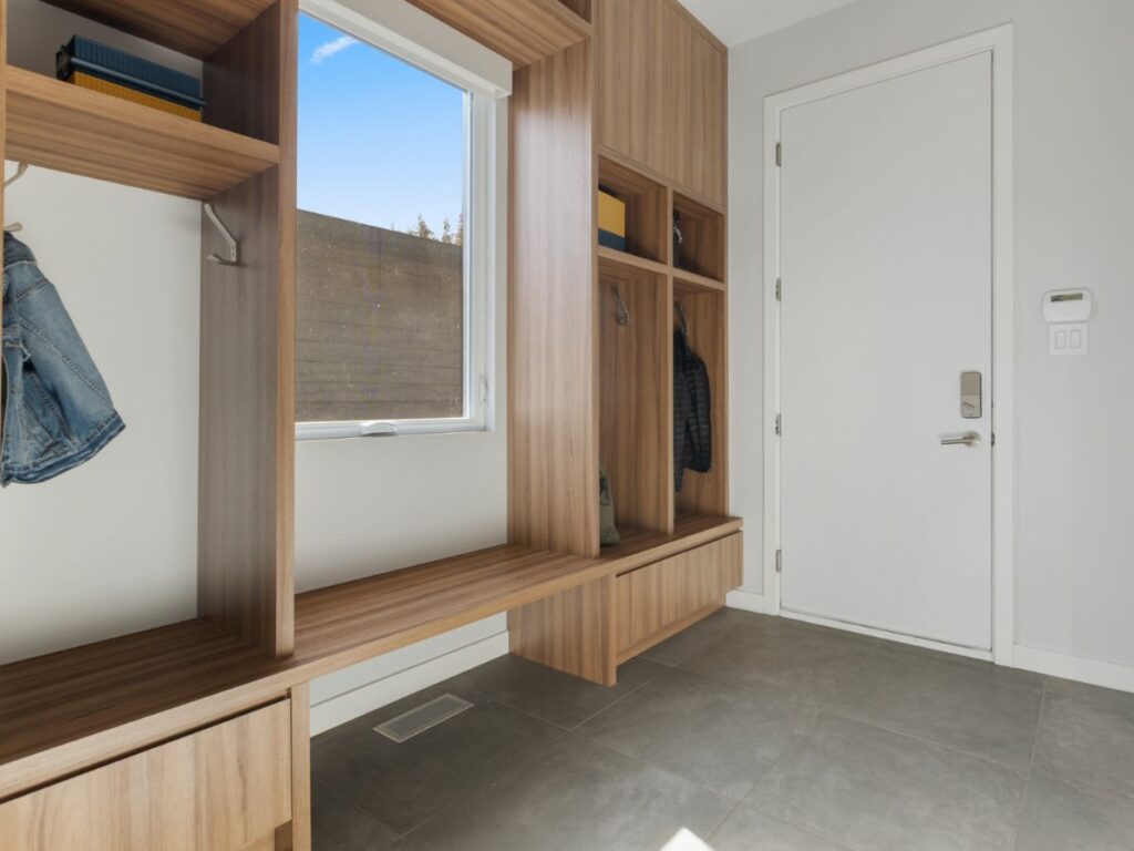 Modern, minimalist entryway with built-in wooden bench and storage shelves against a white door and walls, featuring mudroom organizers.