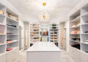 An elegant walk-in closet featuring an array of shelves with shoes and clothing, a central island with a marble countertop, and a crystal chandelier.