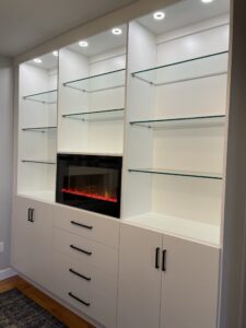 Modern white built-in shelving unit with glass shelves, an electric fireplace, and custom entertainment centers.