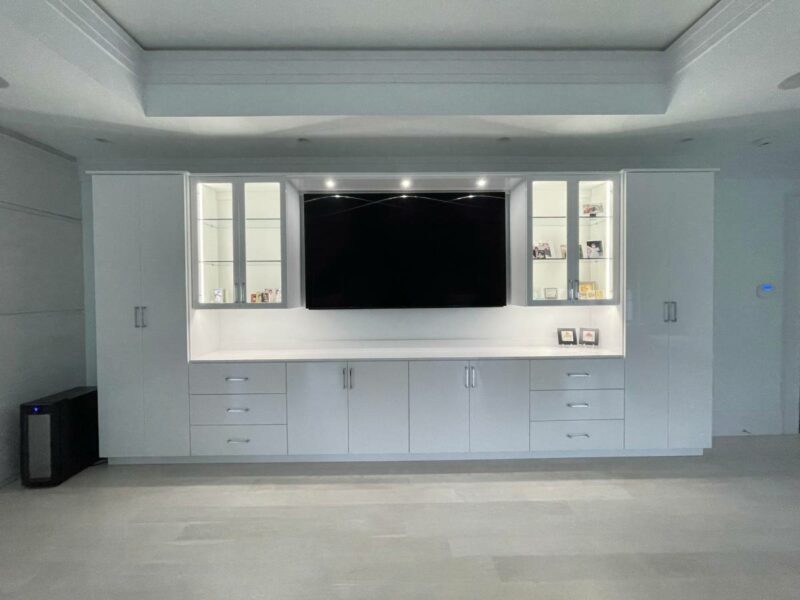 A modern living room featuring a large wall-mounted TV flanked by custom entertainment centers with white shelving units and lower cabinets.