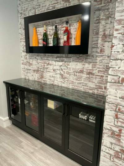 Modern home bar area with custom entertainment centers, featuring glass-fronted cabinets and wall-mounted shelf displaying bottles, against a rustic brick-style wall.