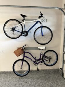 Two bicycles mounted on a wall rack in front of custom garage cabinets, one above the other; the top is a white and blue sports bike and the bottom is a purple cruiser with a basket.
